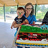 Rj is registered to the contest to win money with this photo: child, toddler, adult, woman, birthday_cake, cake, face_paint, frosting, holding, outdoor, pavilion, celebration, party, toy, fire_truck, glasses, blue_shirt, grass, trees, smile
