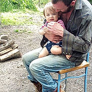 Rj is registered to the contest to win money with this photo: man, baby, child, outdoor, nature, greenery, grass, dirt, wood, firewood, boots, chair, clothing, casual, holding, smiling, affection, portrait, family, parent