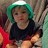 Rj is registered to the contest to win money with this photo: baby, child, hat, green_hat, sitting, couch, blanket, cushion, indoors, cute, infant, foot, hand, clothing, portrait, person, young, comfortable, relaxed, curious