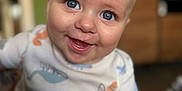 Logan is registered to the contest to win money with this photo: baby, smiling, blue_eyes, onesie, dinosaur_pattern, close_up, infant, happy, child, cute, chubby_hand, indoor, portrait, soft_focus, skin, expression, person, face, young_child, casual