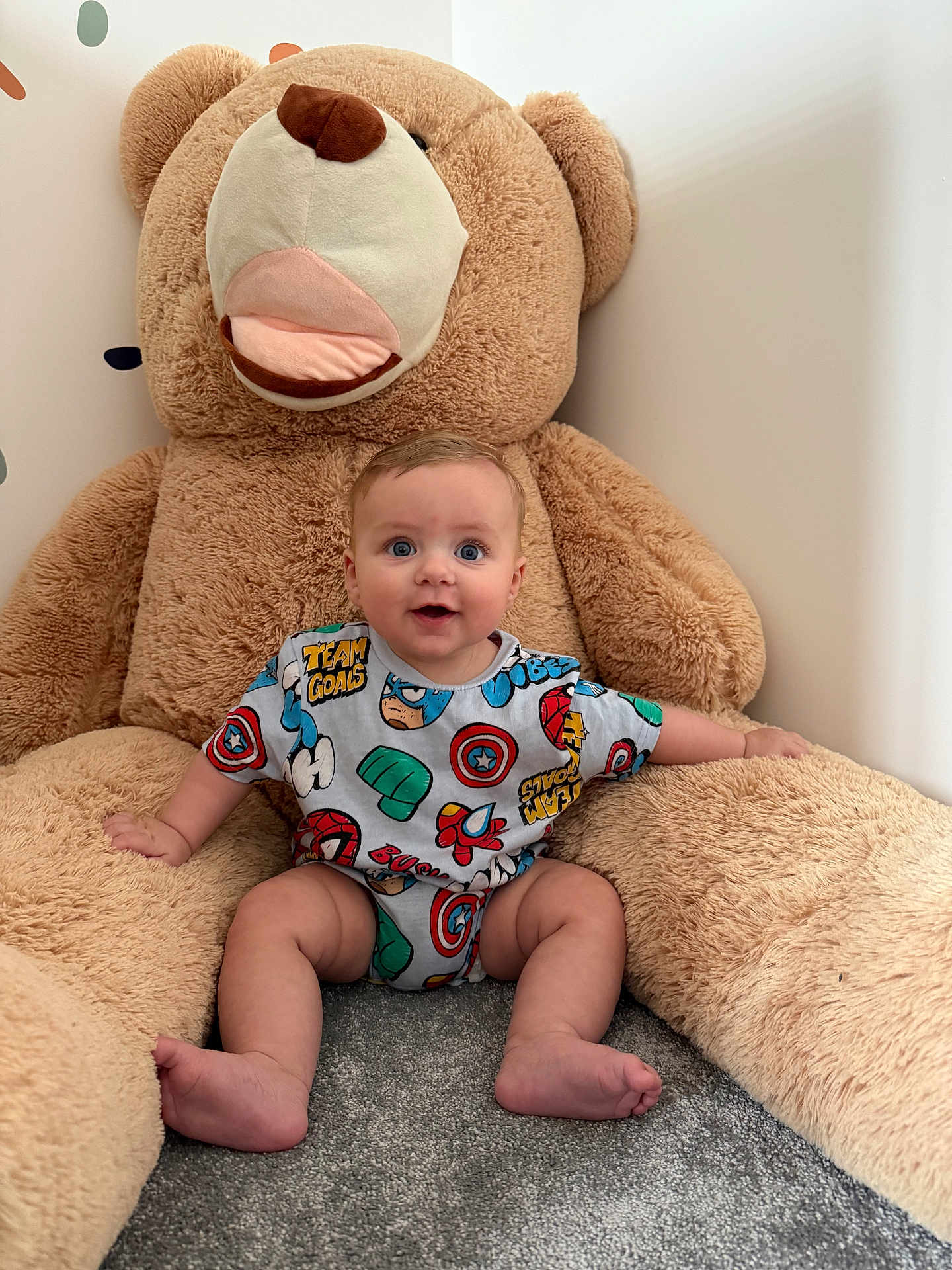 Logan joined the competition — help win amazing prizes! baby, child, teddy_bear, plush_toy, blue_eyes, onesie, superhero_theme, sitting, carpet, smiling, cute, soft_toy, indoors, happy, infant, person, furniture, cuddly, colorful_clothing, young_child