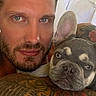 man, dog, french_bulldog, puppy, tattoo, tattooed_arm, bed, white_sheets, close_up, portrait, face, blue_eyes, pet, cuddle, indoor, resting, looking_at_camera, animal, human, companion