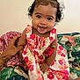 baby, child, smiling, girl, dress, floral_pattern, curly_hair, sitting, indoor, hand, support, colorful, bedspread, happy, portrait, cute, infant, person, young_child, playful
