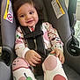 toddler, child, baby, car_seat, onesie, bow, hair_accessory, smile, seatbelt, sitting, indoors, warning_label, safety, cute, infant, person, clothing, hand, face, young_child