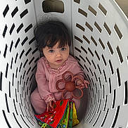 Ava is registered to the contest to win money with this photo: baby, child, laundry_basket, toy, soft_book, flower_toy, pink_clothing, indoor, curly_hair, looking_up, cute, playful, infant, person, sitting, basket, white, floor, colorful, portrait