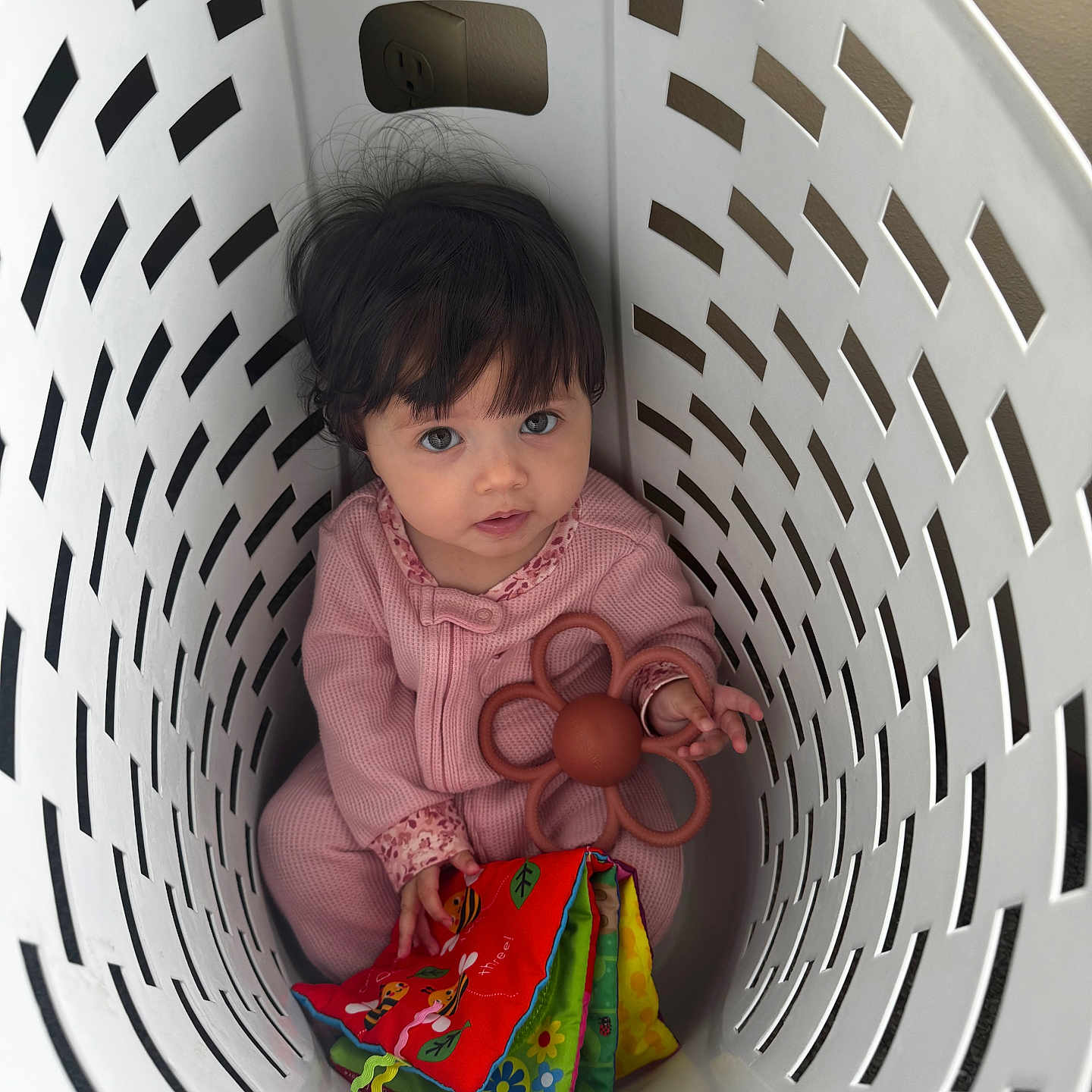 Ava is registered to the contest to win money with this photo: baby, child, laundry_basket, toy, soft_book, flower_toy, pink_clothing, indoor, curly_hair, looking_up, cute, playful, infant, person, sitting, basket, white, floor, colorful, portrait