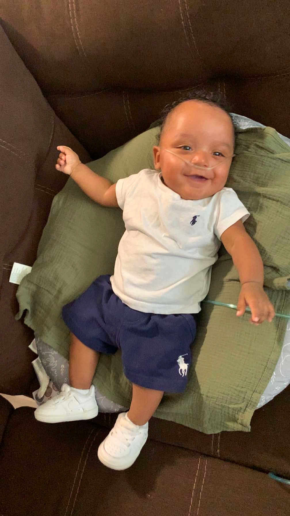 Javien joined the competition — help win amazing prizes! baby, child, smiling, couch, green_cushion, white_tshirt, navy_shorts, white_sneakers, nasal_cannula, happy, indoor, person, infant, clothing, footwear, relaxing, seated, cute, portrait, casual
