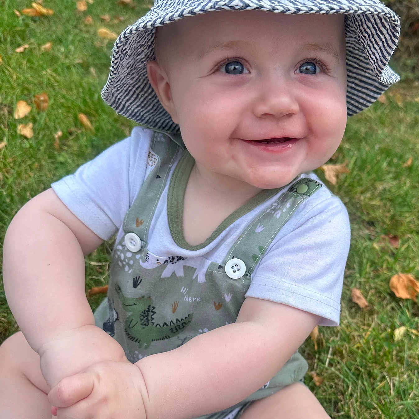 Ollie joined the competition — help win amazing prizes! autumn_leaves, baby, blue_eyes, casual_clothing, child, chubby_hands, cute, dinosaur_print, grass, happy, hat, infant, nature, outdoor, overalls, playful, sitting, smiling, sun_hat, young_child