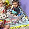 children, toddler, preschooler, baby, faces, serious_expression, playroom, toys, building_blocks, storage_bin, play_mat, indoor, sitting, standing, blond_hair, braided_hair, colorful, family, childcare, portrait