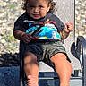 child, toddler, portrait, sitting, chair, outdoor, sunlight, barefoot, curly_hair, smiling, playful, casual_clothing, shorts, graphic_shirt, grass, gravel, fence, shadows, patio, family_photo
