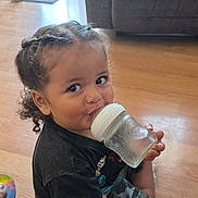 Arlow joined the competition — help win amazing prizes! child, toddler, drinking, baby_bottle, smiling, portrait, sitting, indoors, living_room, wood_floor, sofa, window, book, curly_hair, braids, black_shirt, camouflage_pants, family_home, daylight, happy