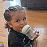 child, toddler, drinking, baby_bottle, smiling, portrait, sitting, indoors, living_room, wood_floor, sofa, window, book, curly_hair, braids, black_shirt, camouflage_pants, family_home, daylight, happy