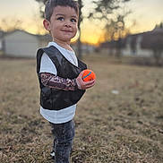 Elias is registered to the contest to win money with this photo: child, boy, basketball, outdoor, sunset, portrait, smiling, standing, grass, field, sports, ball, basketball_player, vest, t_shirt, jeans, sneakers, tattoo_sleeves, trees, residential_background