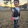 Elias is registered to the contest to win money with this photo: child, toddler, outdoor, portrait, standing, smiling, toy, ball, vest, tshirt, jeans, sneakers, tattoo_sleeve, necklace, grass, yard, trees, sunset, shallow_depth_of_field, golden_hour