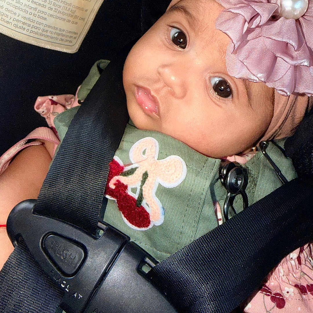 Betsy joined the competition — help win amazing prizes! baby, infant, child, car_seat, safety_harness, pink_headband, flower, pearl, portrait, close_up, wide_eyes, pout, sitting, green_clothing, pink_clothing, indoors, vehicle, securely_strapped, face, photography