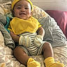 Hezekiah is registered to the contest to win money with this photo: baby, smiling, yellow_clothing, mittens, socks, hat, striped_onesie, cushion, patterned_blanket, indoor, infant, happy, cute, child, person, seated, cozy, soft_lighting, portrait, home