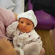 Amelia is registered to the contest to win money with this photo: baby, infant, child, person, clothing, hat, blanket, hand, face, indoor, purple_chair, wooden_floor, cute, newborn, soft_lighting, holding, sitting, looking, white_outfit, cartoon_prints