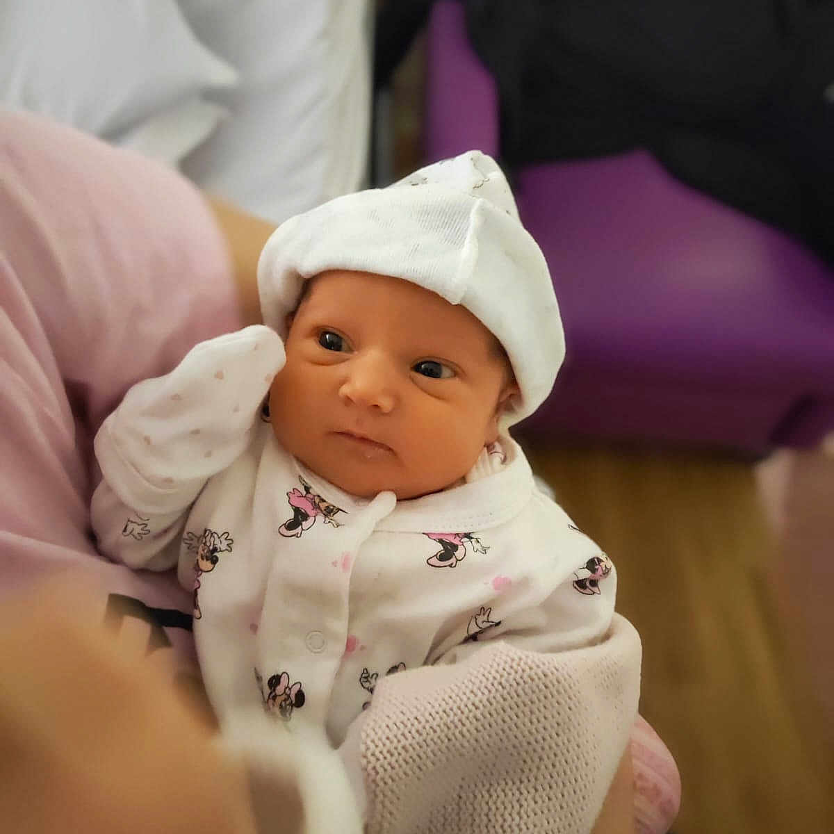 Amelia is registered to the contest to win money with this photo: baby, blanket, cartoon_prints, child, clothing, cute, face, hand, hat, holding, indoor, infant, looking, newborn, person, purple_chair, sitting, soft_lighting, white_outfit, wooden_floor