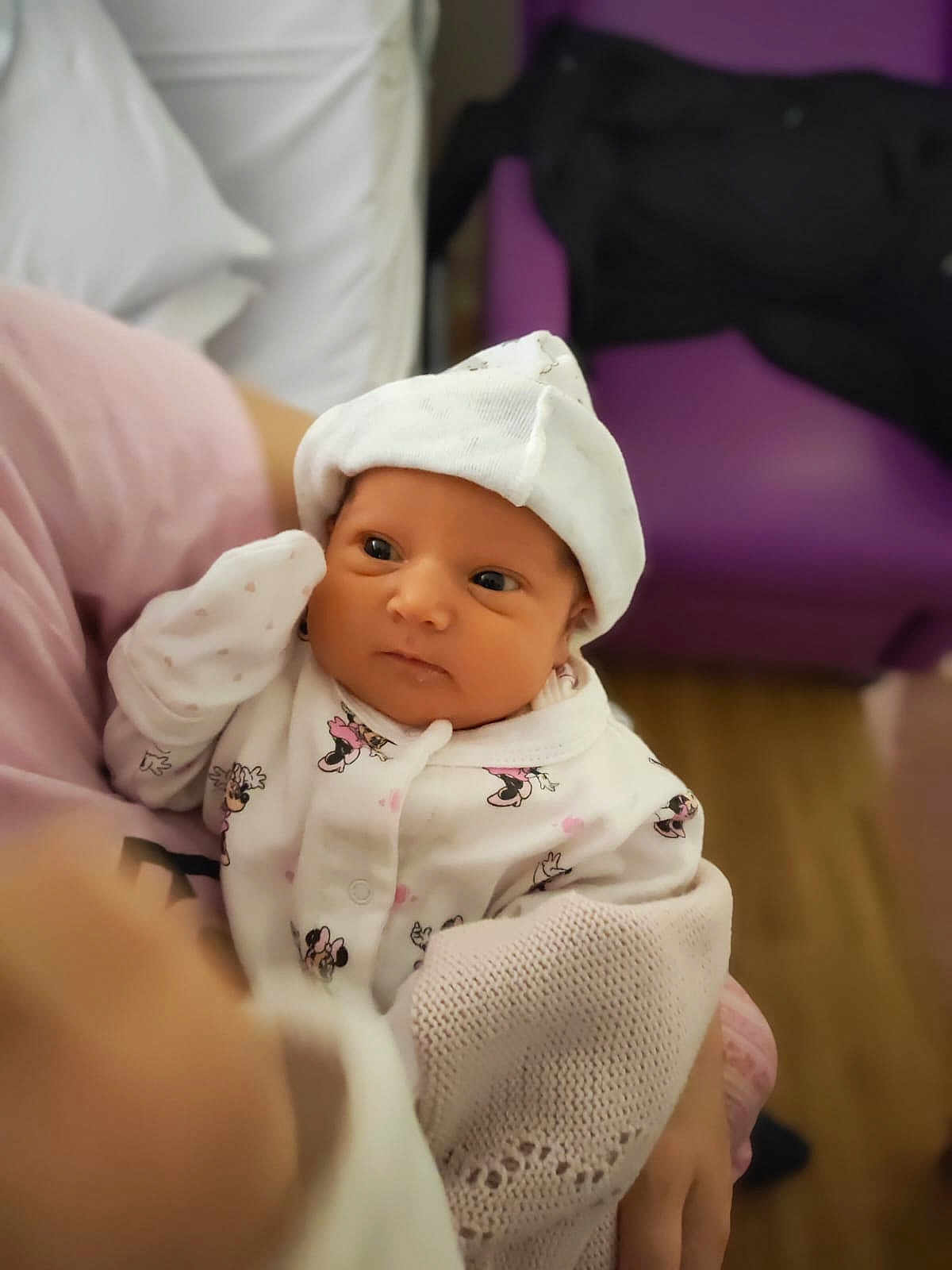 Amelia is registered to the contest to win money with this photo: baby, infant, child, person, clothing, hat, blanket, hand, face, indoor, purple_chair, wooden_floor, cute, newborn, soft_lighting, holding, sitting, looking, white_outfit, cartoon_prints