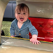 Osiris is registered to the contest to win money with this photo: toddler, child, smiling, blue_sweater, red_cushion, indoor, happy, face, hand, tongue_out, playful, person, young_child, clothing, curly_hair, floor_mat, colorful, cute, portrait, baby