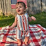 toddler, child, standing, smiling, outdoor, blanket, grass, flag, american_flag, summer, sunlight, striped_clothing, shorts, top, happy, baby, nature, yard, wooden_fence, cute
