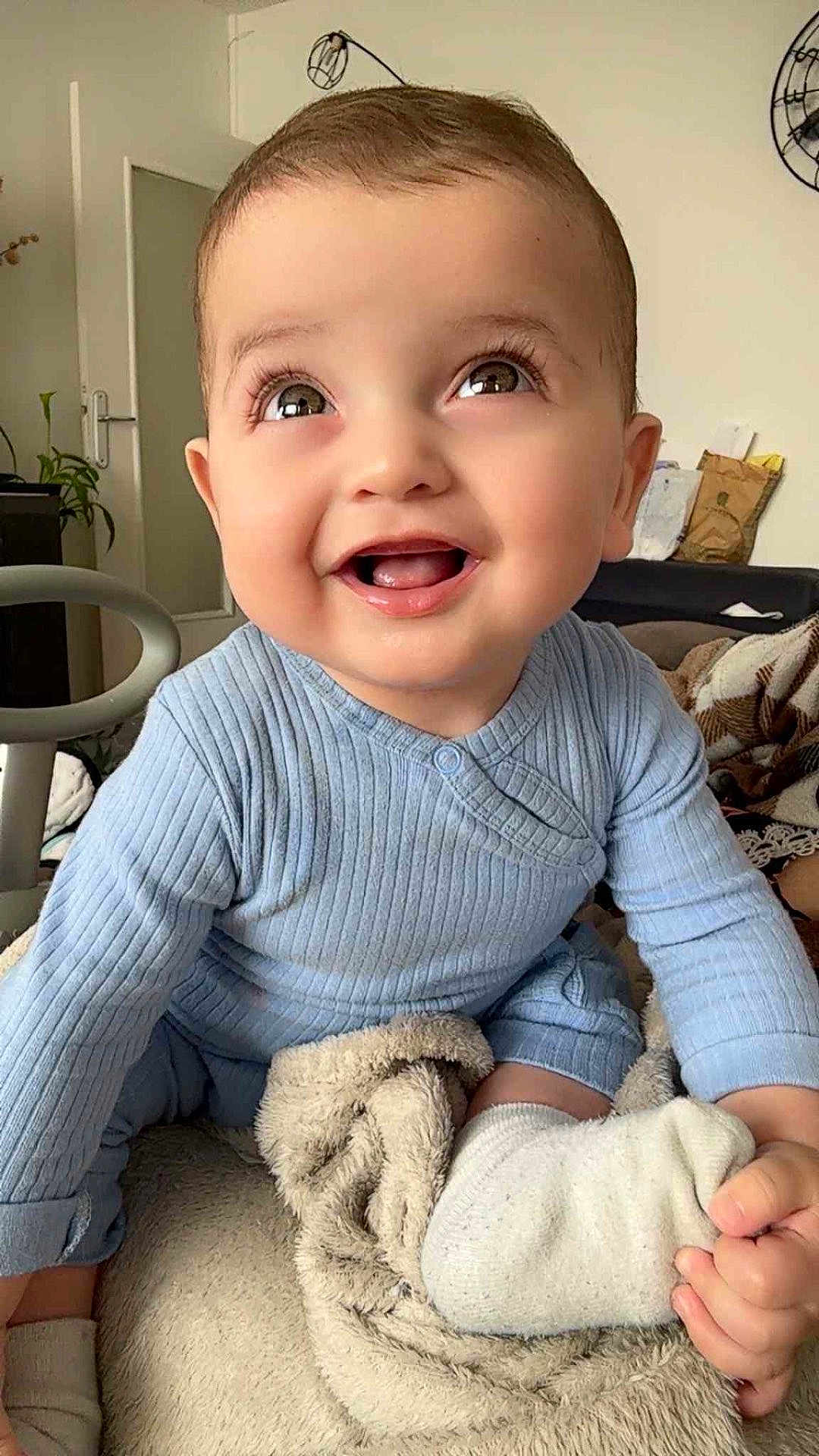 Elyas participe au concours pour gagner de l'argent avec cette photo : baby, smiling, blue_outfit, blanket, indoor, happy, child, cute, face, eyes, hand, sock, furniture, plant, door, living_room, soft_texture, cozy, portrait, infant