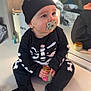 Noah is registered to the contest to win money with this photo: baby, child, pacifier, hat, black_clothing, skeleton_outfit, mirror, reflection, indoor, jar, sitting, table, cute, infant, face, hands, footwear, soft_lighting, expression, room