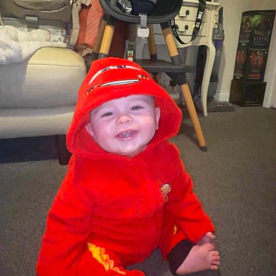 Noah joined the competition — help win amazing prizes! baby, baby_clothing, bottle, carpet, child, couch, floor, footwear_absent, furniture, happy, high_chair, hood, indoor, lamp, living_room, person, red_onesie, smiling, table, toy