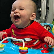 Noah is registered to the contest to win money with this photo: baby, child, laughing, red_shirt, cartoon, toy, activity_center, rings, indoor, washing_machine, happy, play, infant, smile, hand, face, clothing, seat, colorful, fun