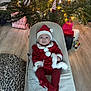 baby, christmas_tree, holiday, red_clothing, santa_hat, festive, indoors, gift, ornament, lights, smile, child, floor, carpet, chair, toy, decoration, warm, cute, portrait
