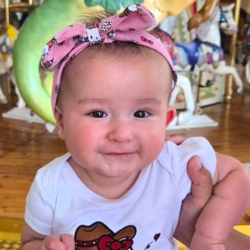 Lilly is registered to the contest to win money with this photo: baby, child, toddler, pink_headband, hello_kitty, carousel, horse, amusement_park, playful, smiling, white_shirt, patterned_pants, indoor, wooden_floor, colorful, fun, person, cute, holding_hand, portrait