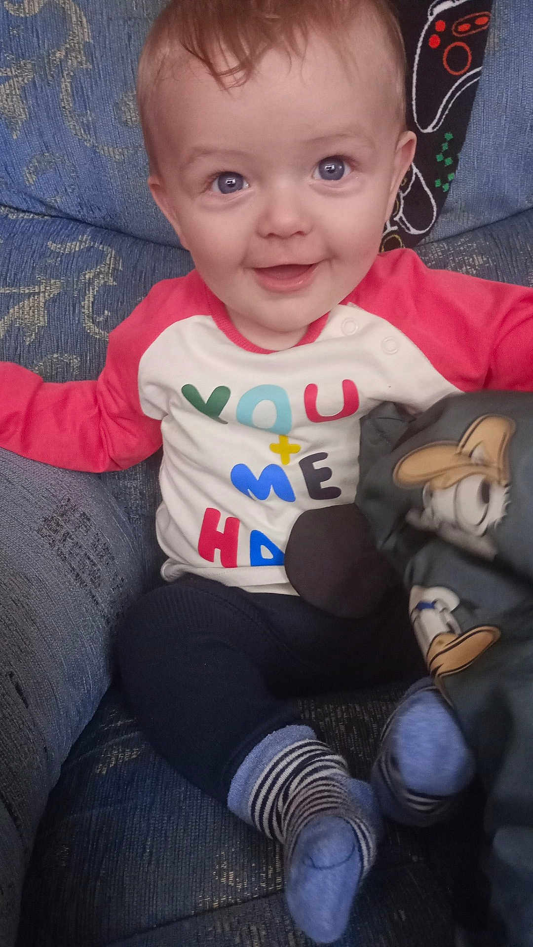 Vincent is registered to the contest to win money with this photo: baby, child, smile, face, blue_eyes, sitting, couch, striped_socks, colorful_shirt, toy, cartoon_character, happy, infant, indoor, clothing, cute, person, young_child, soft_toy, fabric