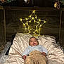baby, infant, smile, blanket, pillow, star_lights, fairy_lights, wood_panel, wooden_frame, garland, ornaments, angel_ornament, bedding, socks, pants, shirt, cozy, holiday, decorations, portrait