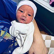 Dean is registered to the contest to win money with this photo: baby, newborn, infant, hat, clothing, person, hand, arm, blue_shirt, indoor, portrait, face, eyes, skin, cozy, blanket, bedroom, toddler, resting, holding