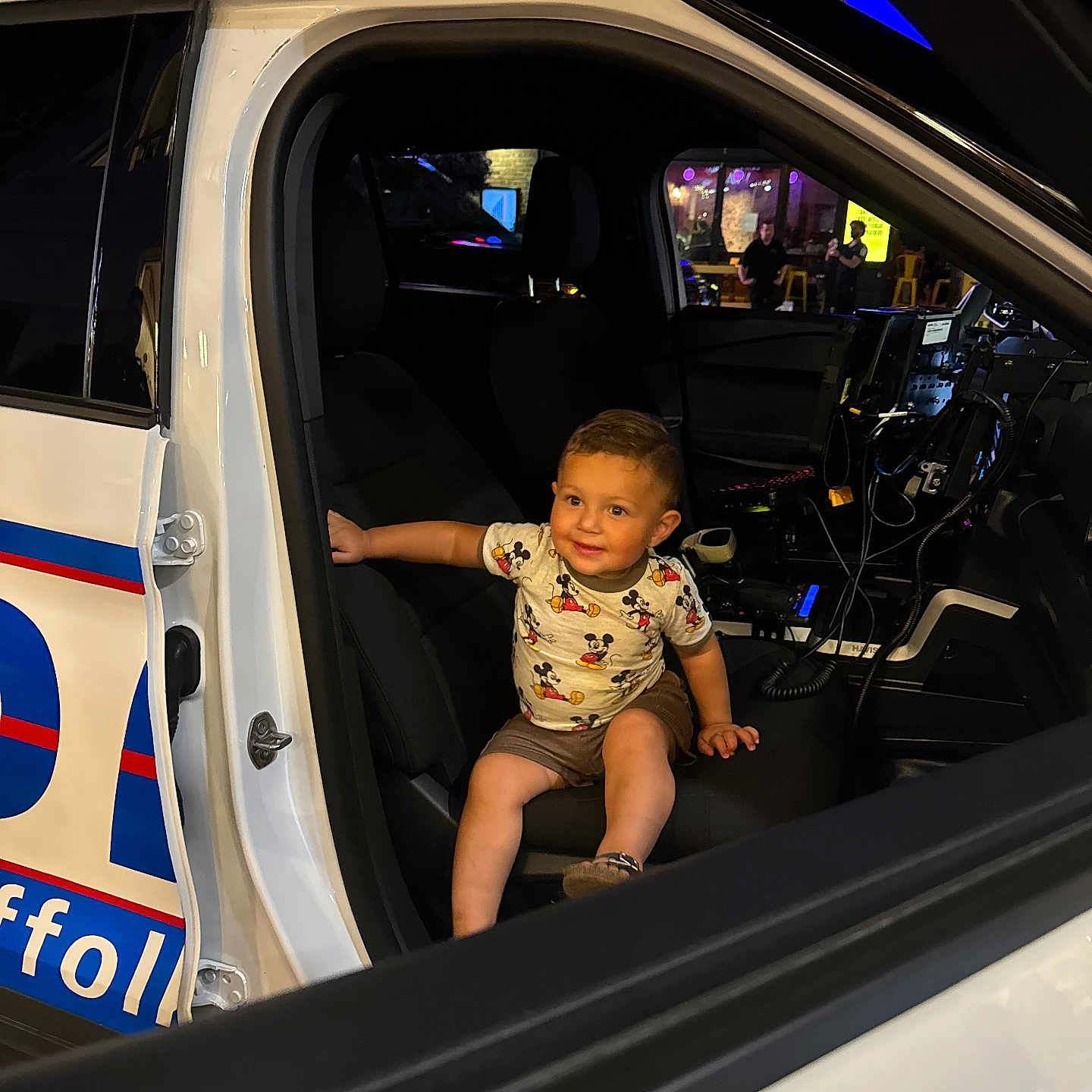 Liam is registered to the contest to win money with this photo: car_interior, child, curious, door, equipment, happy, light, mickey_mouse, night, person, police_car, seat, seatbelt, shirt, shorts, smile, technology, toddler, vehicle, window