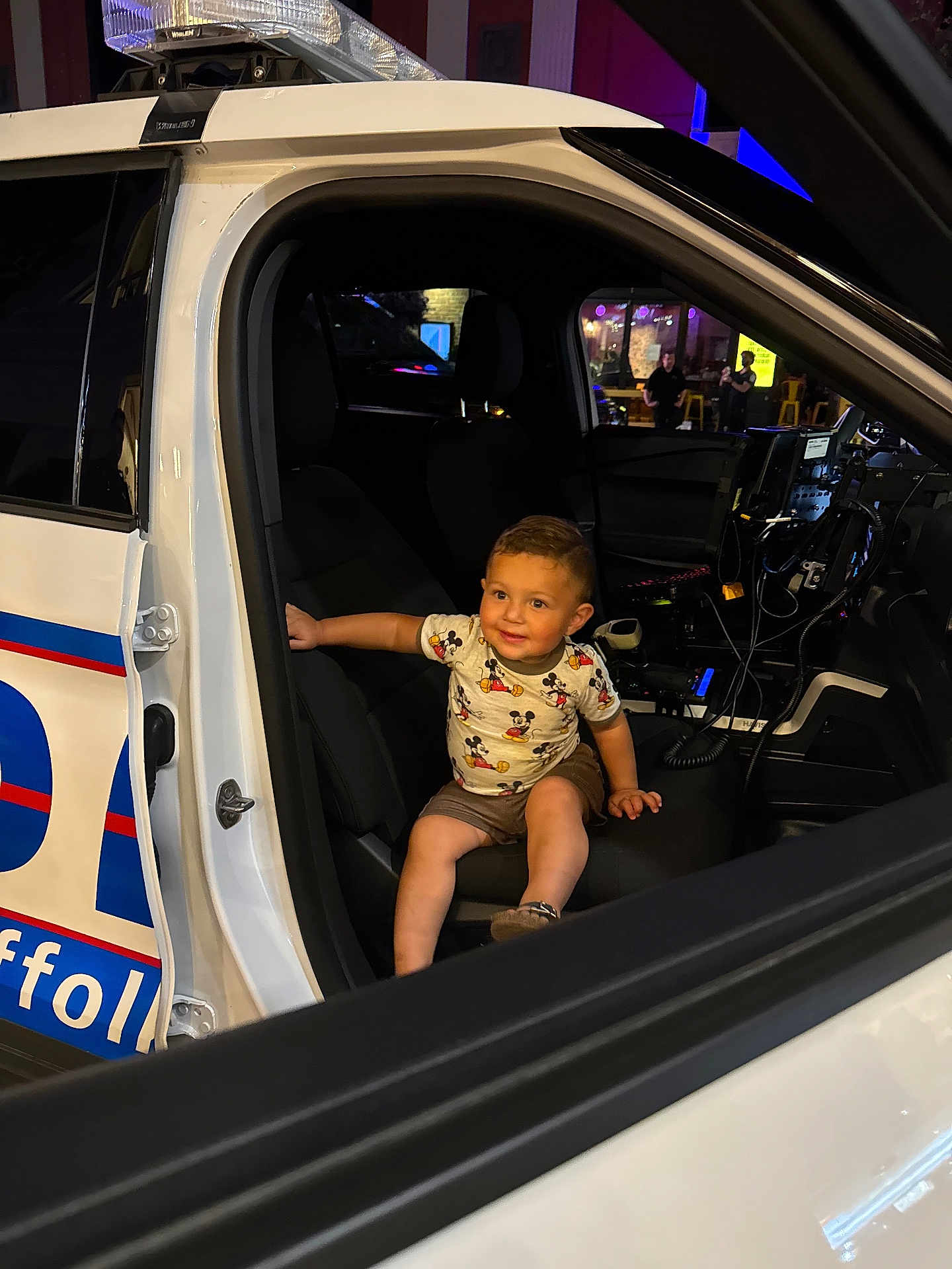 Liam is registered to the contest to win money with this photo: toddler, child, police_car, mickey_mouse, shirt, seat, car_interior, night, smile, shorts, person, technology, vehicle, window, door, equipment, light, seatbelt, happy, curious