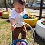 child, toddler, easter_egg, basket, outdoor, grass, toy, spring, colorful, egg_hunt, shiny, curious, pants, white_shirt, sunlight, decor, flower, play, daytime, activity