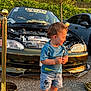 Liam is registered to the contest to win money with this photo: toddler, child, car, hood_open, sunlight, outdoor, parking_lot, shorts, blue_shirt, mickey_mouse, shoes, checkered, golden_hour, vehicle, happy, standing, smiling, engine, car_meet, pole
