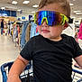toddler, child, shopping_cart, sunglasses, black_shirt, clothing_store, retail, shopping, reflection, casual_wear, indoor, person, fashion, purple_pants, seatbelt, young_child, store_aisle, rack, apparel, modern
