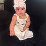 baby, child, headband, bow, white_clothing, sitting, chair, black_chair, indoor, cute, portrait, infant, cute_expression, plush, cozy, small_bracelet, sock, curious, wide_eyes, pouting
