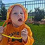 Seven is registered to the contest to win money with this photo: baby, casual_clothing, child, daylight, expression, fence, grass, greenery, hands, mouth_open, nature, orange_shirt, outdoor, playful, sitting, summer, sunlight, toddler, toy, yellow_hat