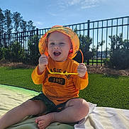Seven is registered to the contest to win money with this photo: baby, blanket, casual_clothing, child, daylight, fence, grass, greenery, happy, nature, outdoor, playful, portrait, shorts, smiling, summer, sunglasses, sunhat, sunny, toddler