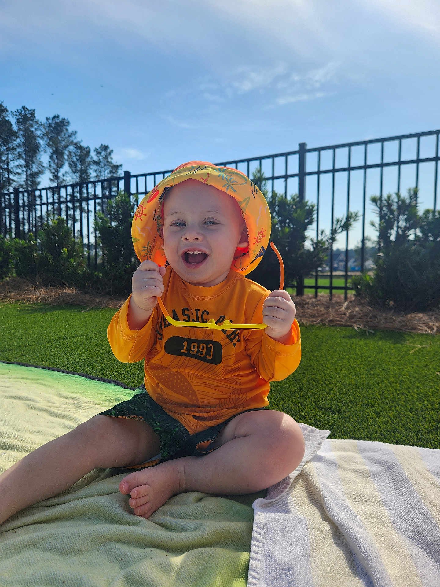 Seven is registered to the contest to win money with this photo: baby, blanket, casual_clothing, child, daylight, fence, grass, greenery, happy, nature, outdoor, playful, portrait, shorts, smiling, summer, sunglasses, sunhat, sunny, toddler