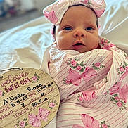 Anaila is registered to the contest to win money with this photo: baby, newborn, infant, blanket, headband, pink, floral, bed, plaque, birth_details, handwritten, cute, wrapped, portrait, sleeping_area, soft, closeup, person, child, indoors