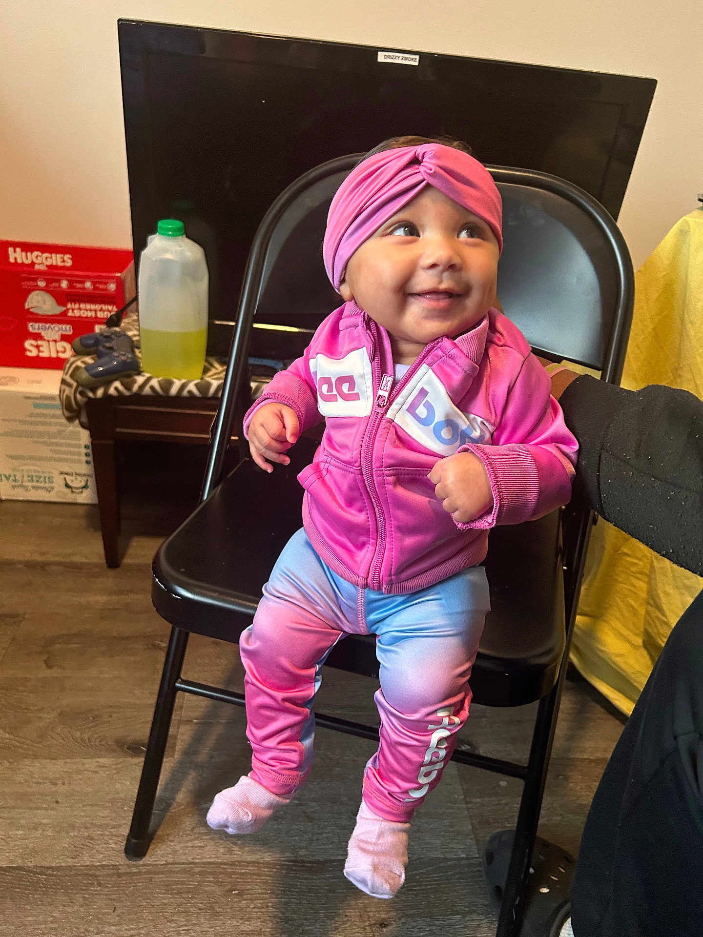 Laveah joined the competition — help win amazing prizes! baby, child, smiling, pink_clothing, headband, sitting, chair, indoor, floor, person, hand, television, bottle, box, wooden_floor, casual, happy, cute, support, pants