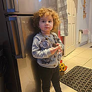 Kataleya joined the competition — help win amazing prizes! toddler, child, curly_hair, barefoot, sweater, necklace, kitchen, refrigerator, tile_floor, rug, door, towel, wall_decor, toy, earrings, standing, indoor, expression, person, home