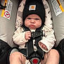 baby, infant, car_seat, black_beanie, clothing, straps, buckle, cute, expression, face, person, child, legs, hand, sitting, indoors, warning_labels, fabric, safety, seat