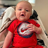 Kashtin is registered to the contest to win money with this photo: baby, infant, child, red_clothing, baseball_graphic, blanket, sitting, indoors, portrait, cute, face, hand, short_sleeves, shorts, person, young_child, cozy, expression, looking_at_camera, comfortable