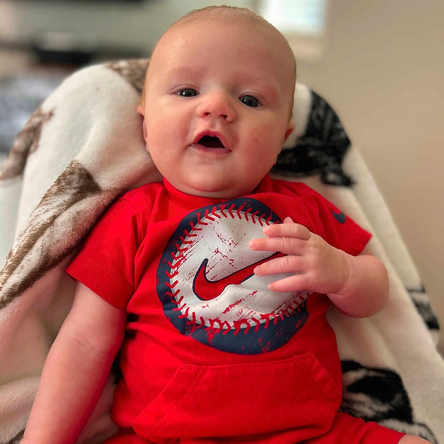 Kashtin is registered to the contest to win money with this photo: baby, baseball_graphic, blanket, child, comfortable, cozy, cute, expression, face, hand, indoors, infant, looking_at_camera, person, portrait, red_clothing, short_sleeves, shorts, sitting, young_child