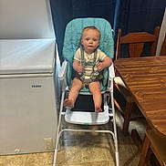 Colton is registered to the contest to win money with this photo: toddler, child, high_chair, baby, indoor, kitchen, table, chair, curtains, floor, freezer, wooden_furniture, straps, person, face, expression, footwearless, clothing, seat, home